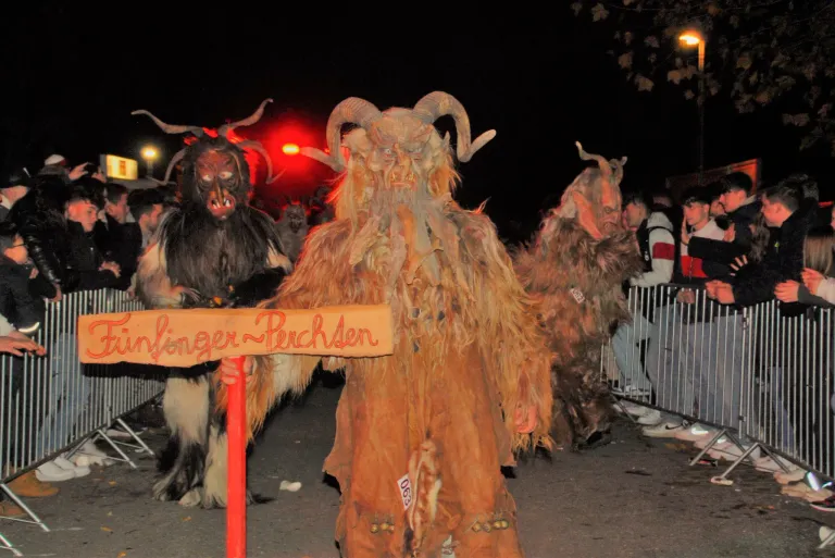 A group of people in horned costumes standing in a parade at night. One holds a sign with red writing. A crowd is watching behind a metal barrier.