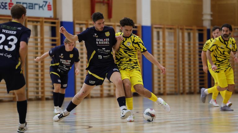 Drei Fußballspieler auf einem Hallenplatz, einer in einem blauen und schwarzen Trikot, ein anderer in einem gelben und schwarzen Tiger-Print-Trikot und ein Dritter im Hintergrund in einem blauen Trikot. Sie sind aktiv an einem Fußballspiel beteiligt.