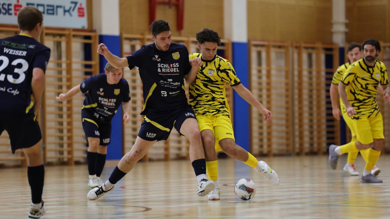 Drei Fußballspieler auf einem Hallenplatz, einer in einem blauen und schwarzen Trikot, ein anderer in einem gelben und schwarzen Tiger-Print-Trikot und ein Dritter im Hintergrund in einem blauen Trikot. Sie sind aktiv an einem Fußballspiel beteiligt.