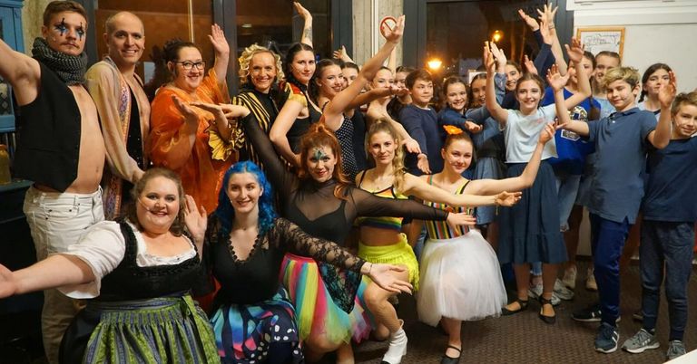 A group of people dressed in various costumes are posing for a photo, some with hands raised and others with painted faces. They are standing and sitting on a tiled floor, with some wearing shoes and others barefoot.