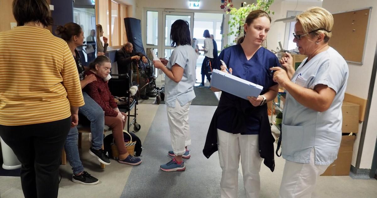 Eine Gruppe von Menschen in einem Krankenhausflur. Eine Krankenschwester hält eine Mappe und einen Stift und spricht mit einer Patientin im Rollstuhl. Ein anderer Patient sitzt auf einer Bank, während andere vorbeigehen.