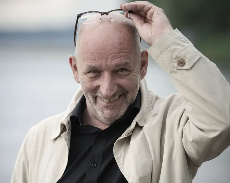 Ein kahlköpfiger Mann in einem schwarzen Hemd und einer beigen Jacke lächelt und hält seine Brille auf dem Kopf.