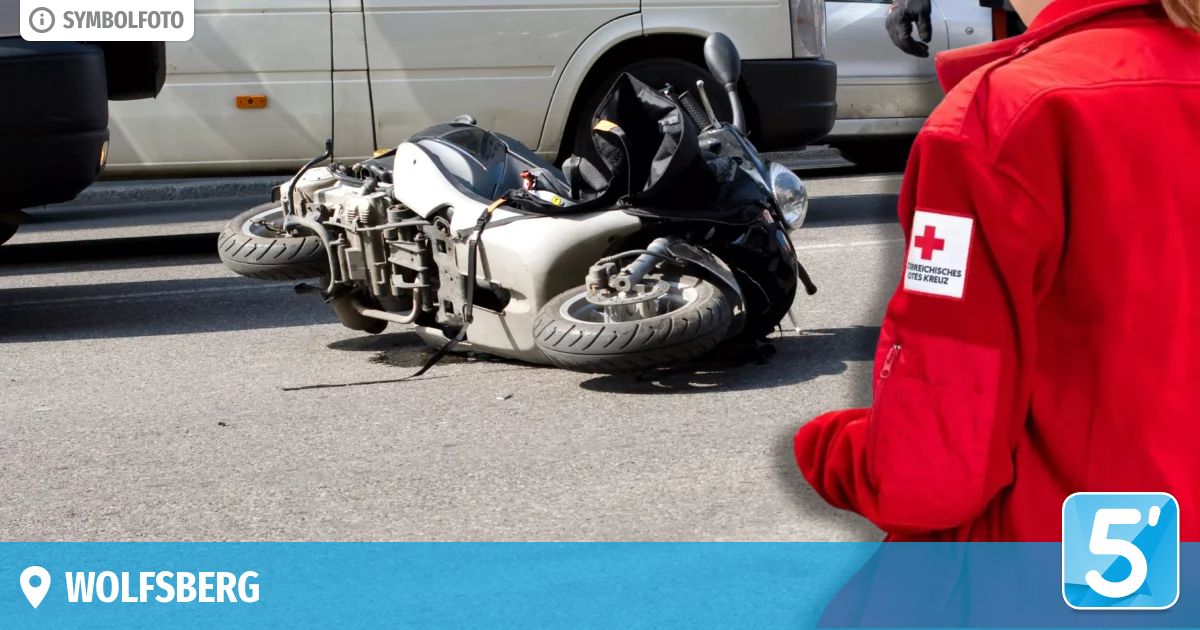 Ein Motorrad liegt mit abgerissenem Vorderrad auf der Straße. Eine Person in einem roten Mantel mit rotem Kreuz-Patch steht in der Nähe. Ein weißer Van ist hinter dem Motorrad geparkt.