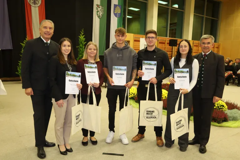 Eine Gruppe von fünf Personen in formeller Kleidung steht und hält Urkunden und Taschen, während sie für ein Foto lächeln. Sie stehen vor einem Hintergrund mit Fahnen und einer Holzstruktur.