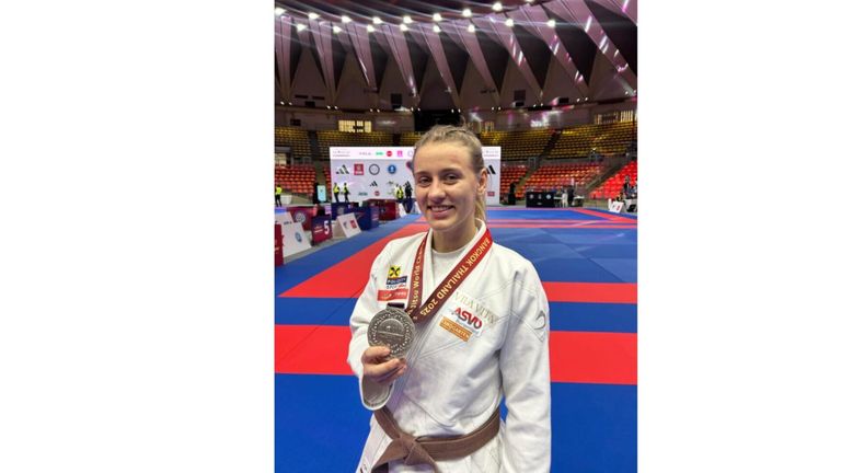 Eine Frau in einem weißen Kampfsportanzug mit einer Silbermedaille um den Hals steht auf einem Teppich in einer überdachten Sportarena. Die Arena hat einen roten und blauen Boden, und es gibt Banner mit Logos und Text im Hintergrund.