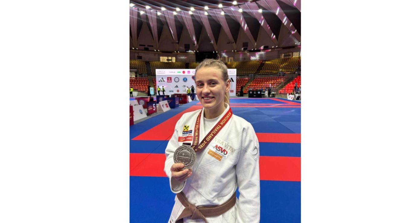 Eine Frau in einem weißen Kampfsportanzug mit einer Silbermedaille um den Hals steht auf einem Teppich in einer überdachten Sportarena. Die Arena hat einen roten und blauen Boden, und es gibt Banner mit Logos und Text im Hintergrund.