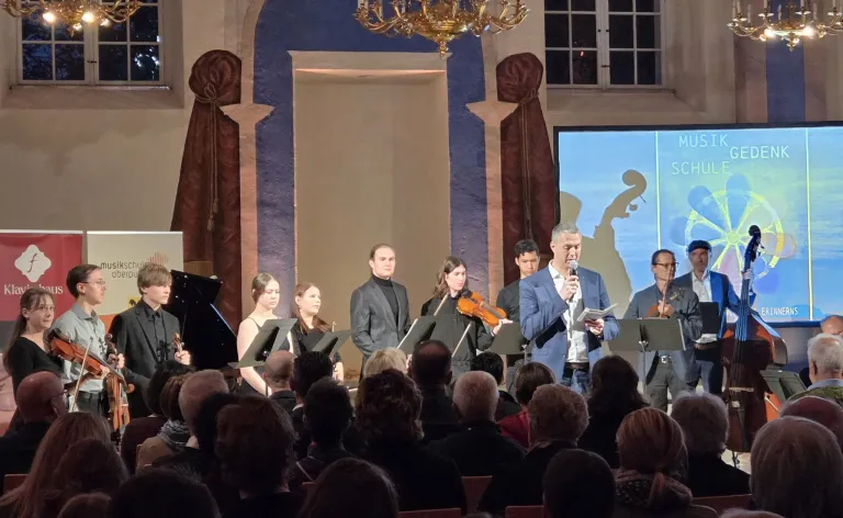 Ein klassisches Musikensemble tritt auf der Bühne auf, mit Geiger, Pianist und Dirigent. Ein Publikum sitzt davor und beobachtet eine große Leinwand hinter den Musikern.