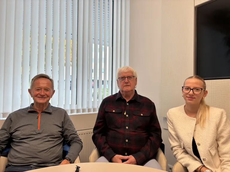 Three individuals are seated in an office setting, smiling for a photo. A man with glasses and a checkered shirt is in the middle, with a smiling man in a gray jacket on his left and a woman in a white coat on his right. Behind them is a window with blinds.