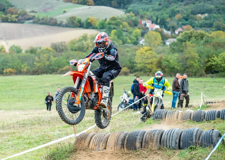 Ein Motocross-Fahrer springt über gestapelte Reifen auf einer Erdpiste. Zuschauer beobachten von den Seitenlinien. Der Hintergrund zeigt eine üppige, grüne Landschaft mit Hügeln.
