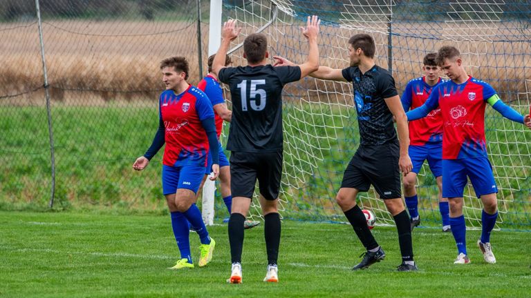 Fußballspieler feiern ein Tor mit High-Fives vor einem Netz, tragen Teamtrikots mit Nummern und Marken.
