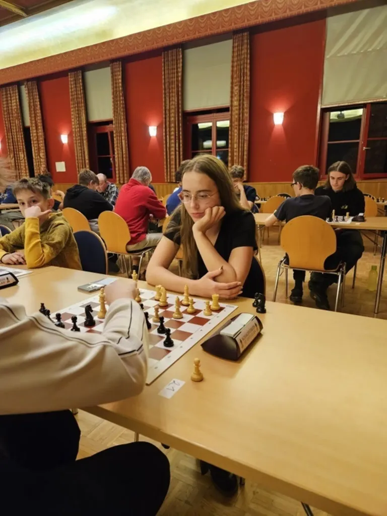 Ein Schachturnier findet in einem Raum mit roten Wänden und Holzstühlen statt. Eine Frau mit Brille konzentriert sich auf das Spiel vor ihr.