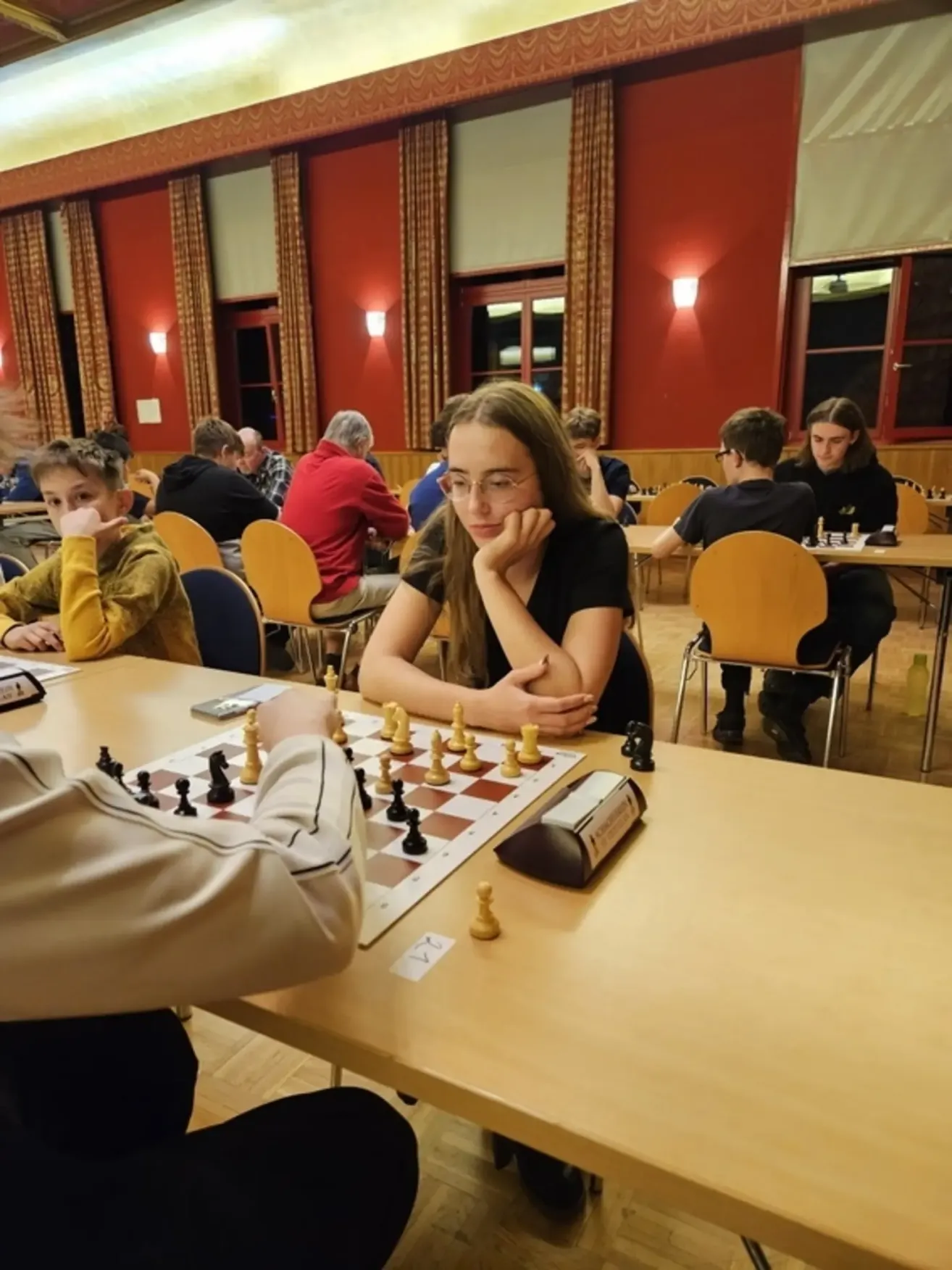 Ein Schachturnier findet in einem Raum mit roten Wänden und Holzstühlen statt. Eine Frau mit Brille konzentriert sich auf das Spiel vor ihr.