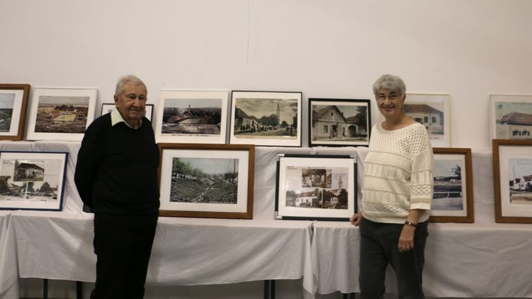 Ein älterer Mann und eine ältere Frau stehen neben einem Tisch mit gerahmten Fotografien alter Gebäude und Landschaften. Sie lächeln und scheinen sich zu amüsieren.