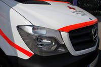 Ein weißes und rotes Mercedes-Benz-Fahrzeug, vermutlich ein Krankenwagen, zeigt ein rotes Kreuz-Symbol auf der Motorhaube und hat ein Nummernschild unten rechts.