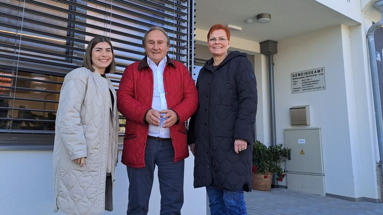 Three people stand outside a building. A man in a red jacket, a woman in a white coat, and another woman in a black jacket. They are smiling and posing for a photo.