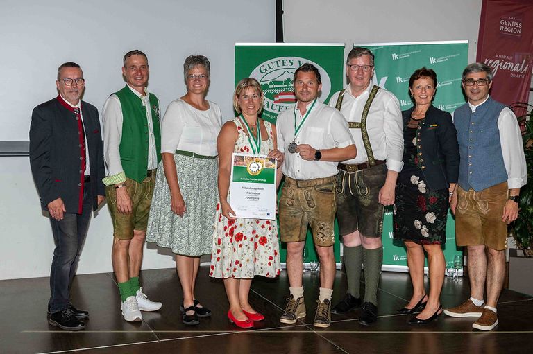 Eine Gruppe von Menschen in traditioneller deutscher Kleidung, einschließlich Lederhosen und Dirndln, steht zusammen. Eine Frau in der Mitte hält ein Zertifikat und eine Medaille. Sie posieren für ein Foto vor einem grünen Banner mit Text.