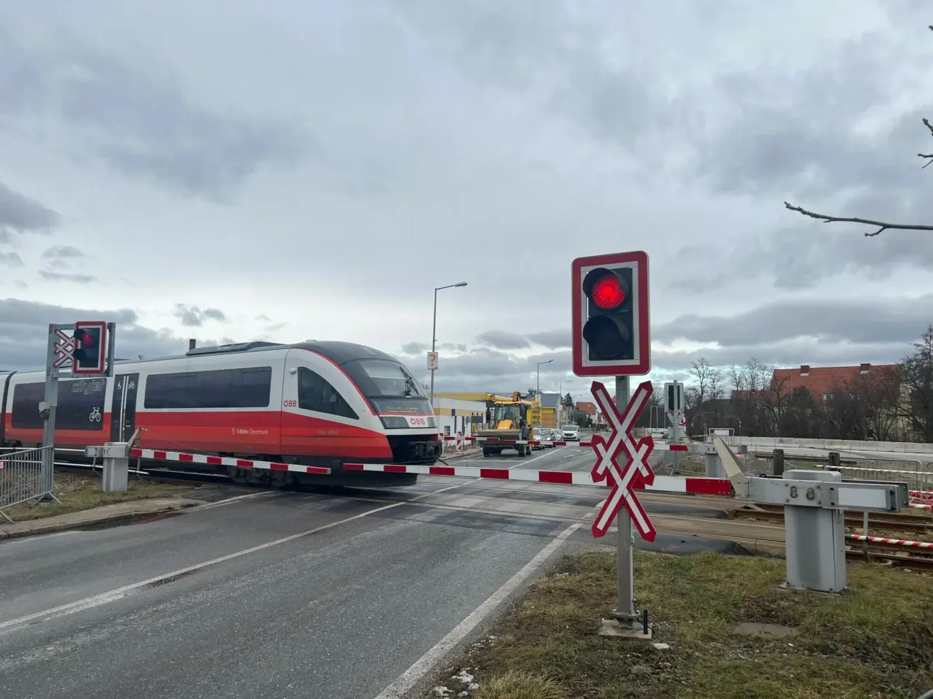 A train is stopped at a railroad crossing with a red light. The road is closed with barriers, and a construction vehicle is nearby.