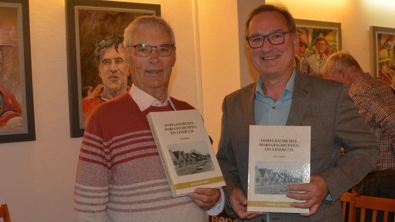 Zwei Männer lächeln und halten Bücher, möglicherweise bei einer Buchsignierstunde. Der Mann auf der linken Seite trägt einen roten Pullover, während der Mann auf der rechten Seite einen grauen Anzug trägt. Sie tragen beide eine Brille.