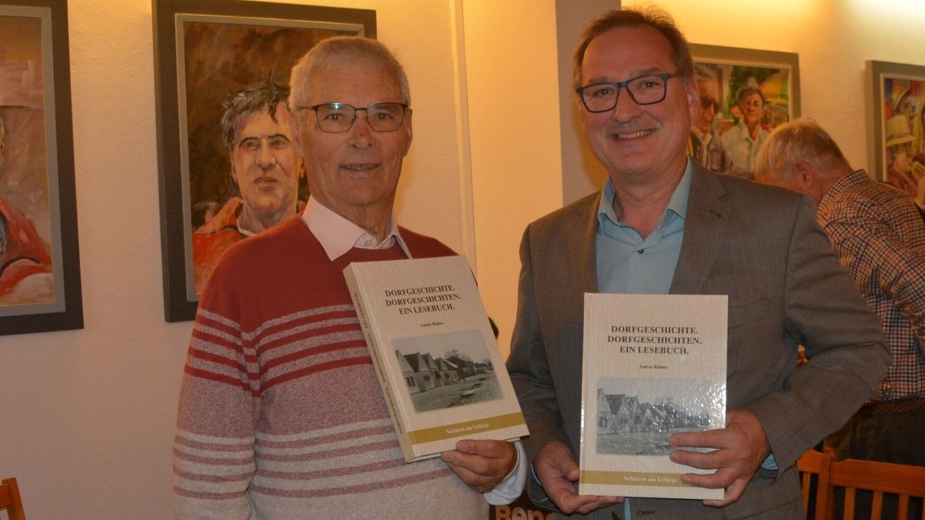 Zwei Männer lächeln und halten Bücher, möglicherweise bei einer Buchsignierstunde. Der Mann auf der linken Seite trägt einen roten Pullover, während der Mann auf der rechten Seite einen grauen Anzug trägt. Sie tragen beide eine Brille.