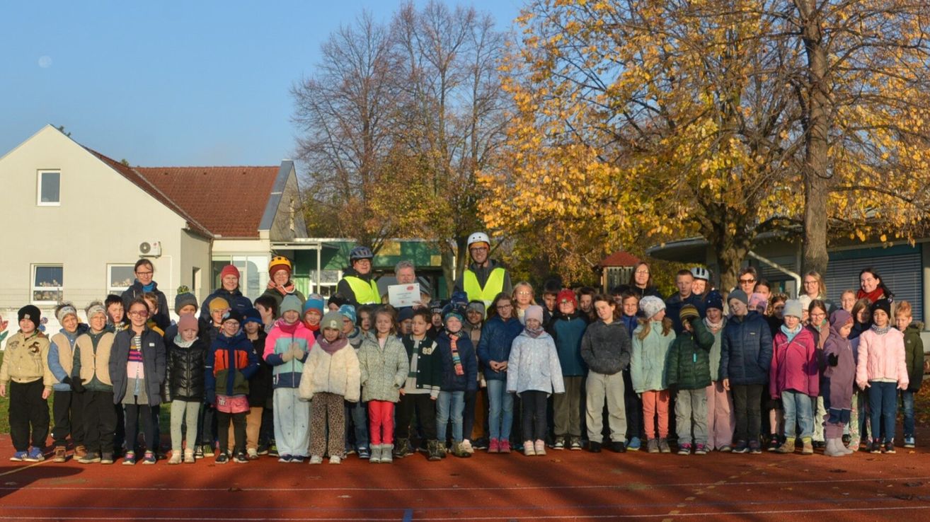 Eine Gruppe von Kindern und Erwachsenen steht auf einer Laufbahn mit einem Zertifikat, trägt Winterkleidung und Helme. Bäume und Gebäude sind im Hintergrund.