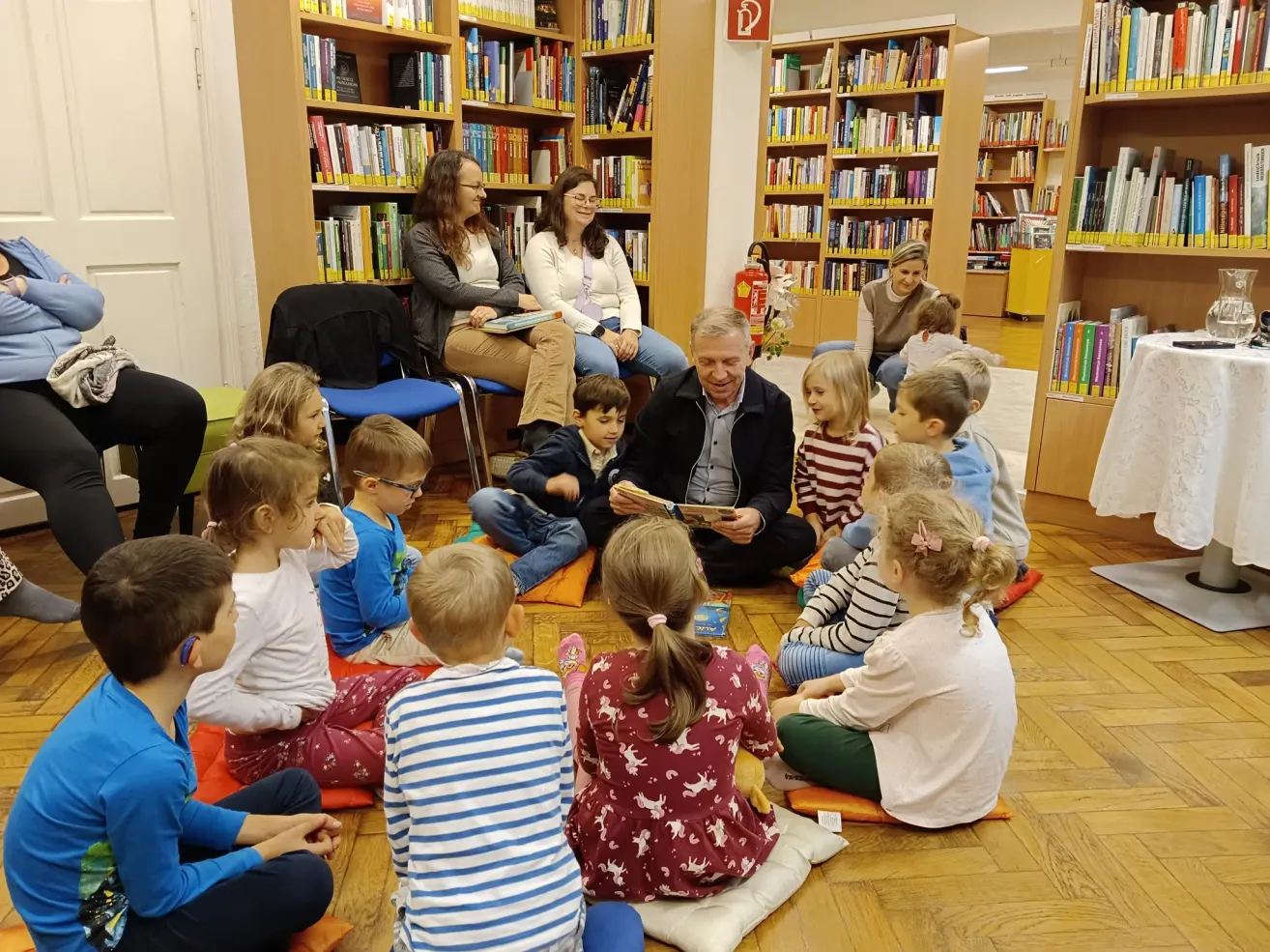 A group of children and adults are sitting in a circle in a library. A man reads a book while others listen. Shelves filled with books are in the background.