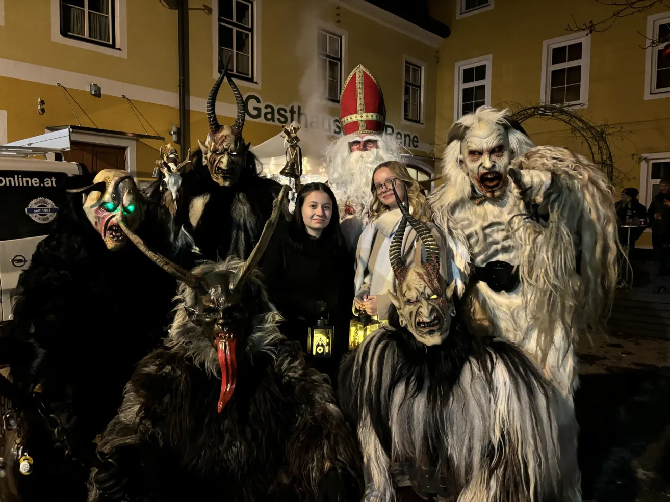 A group of people dressed in scary costumes are posing for a photo in front of a building. A girl is holding a lantern.