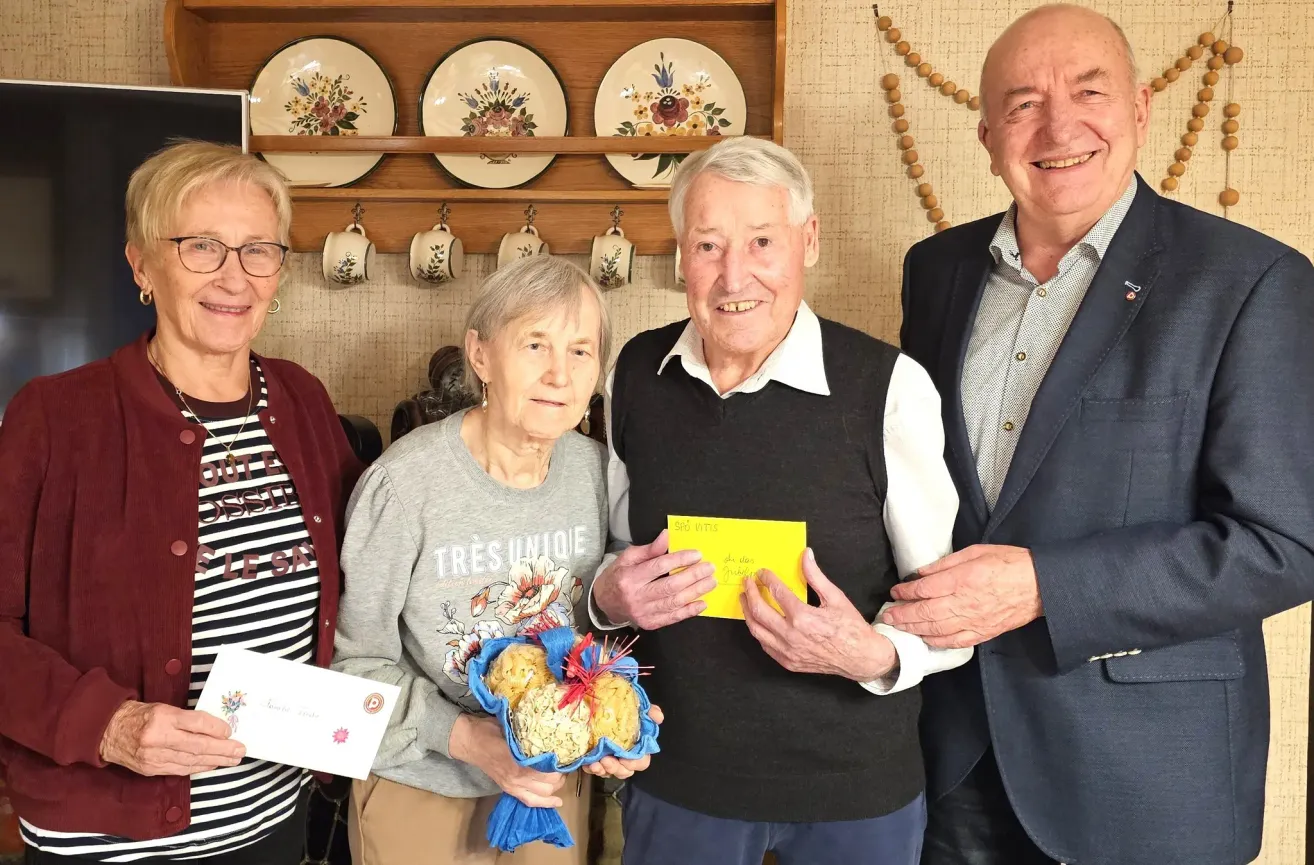 Four elderly people stand in a room. Two men and two women. The women are smiling and holding a bouquet of food. The men are smiling too. They are holding a yellow paper. The room has a wooden shelf with plates and cups.