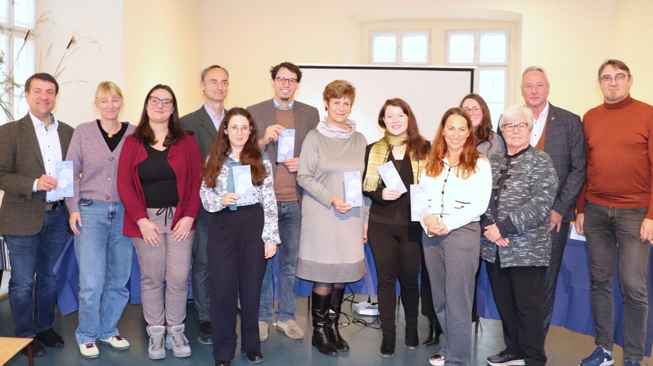 Eine Gruppe von Personen posiert für ein Foto. Sie halten Bücher in den Händen, und einige tragen Brillen. Hinter ihnen befindet sich eine Tafel und ein Fenster.