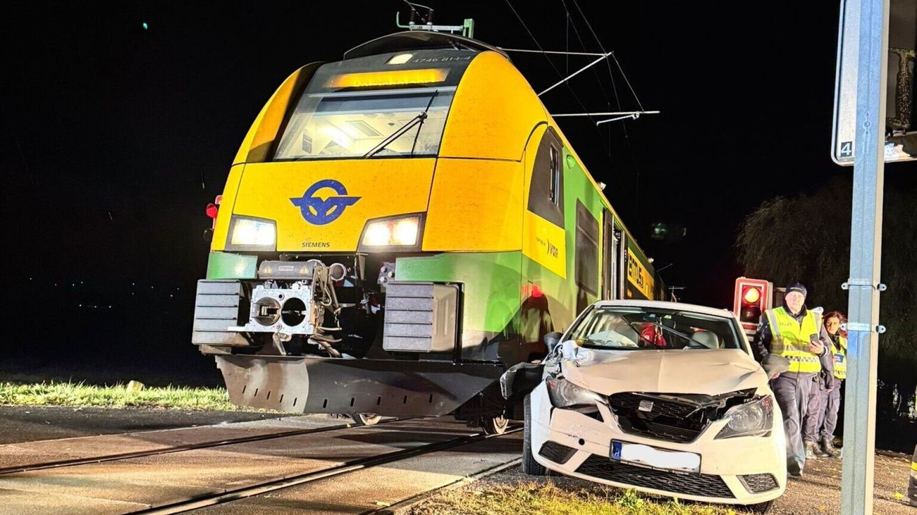 A yellow and green train with a blue logo and the text Siemens is parked on a track. Next to it, a white car with a damaged front is on the grass.