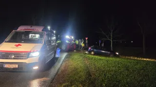 At night, an ambulance is parked on the road with a tractor and emergency workers standing nearby. A blue car is wrecked on the side of the road.
