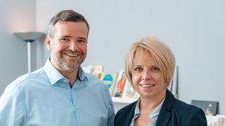 A man and a woman, both smiling, stand next to each other in an office. Behind them, a bookshelf holds books and photos.