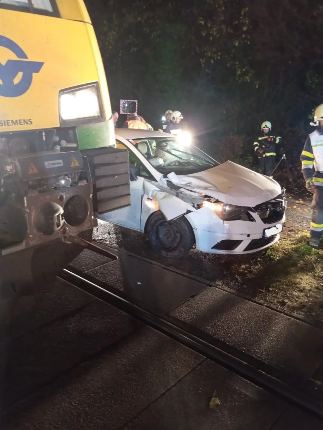 A white car has collided with a train on a track. Emergency responders in yellow and black uniforms are attending to the scene.