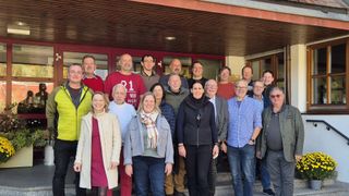 A group of people in winter attire stand on steps in front of a building, smiling for a group photo. They are diverse in age and gender, with some wearing glasses and scarves. The background features glass doors with red frames.