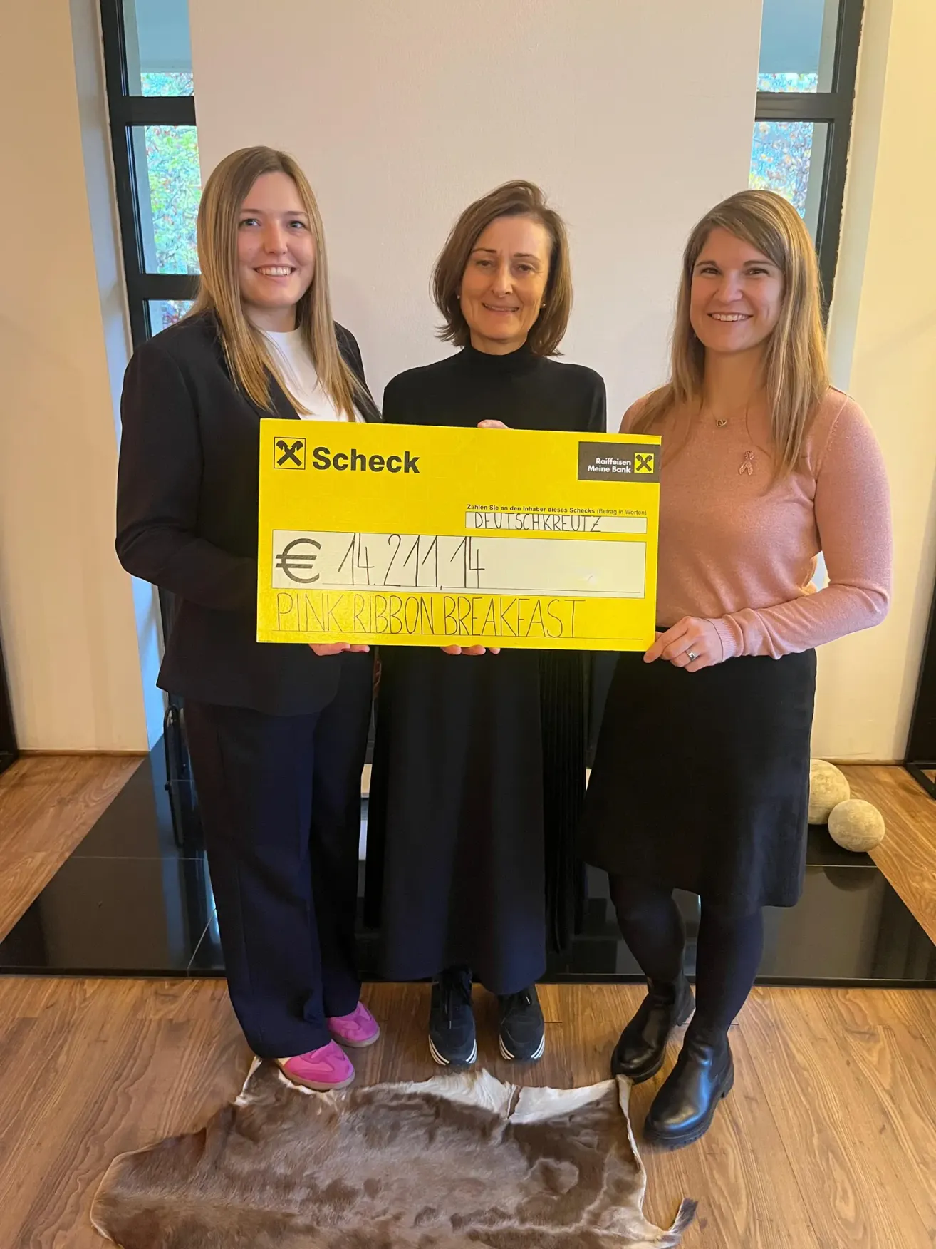 Three women holding a large cheque for 14,241.14 euros, labeled Pink Ribbon Breakfast, standing in a room with a fireplace and wooden floors.