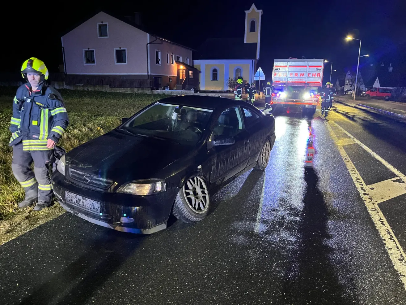 A black car with a flat tire is parked on a road at night with firefighters around.
