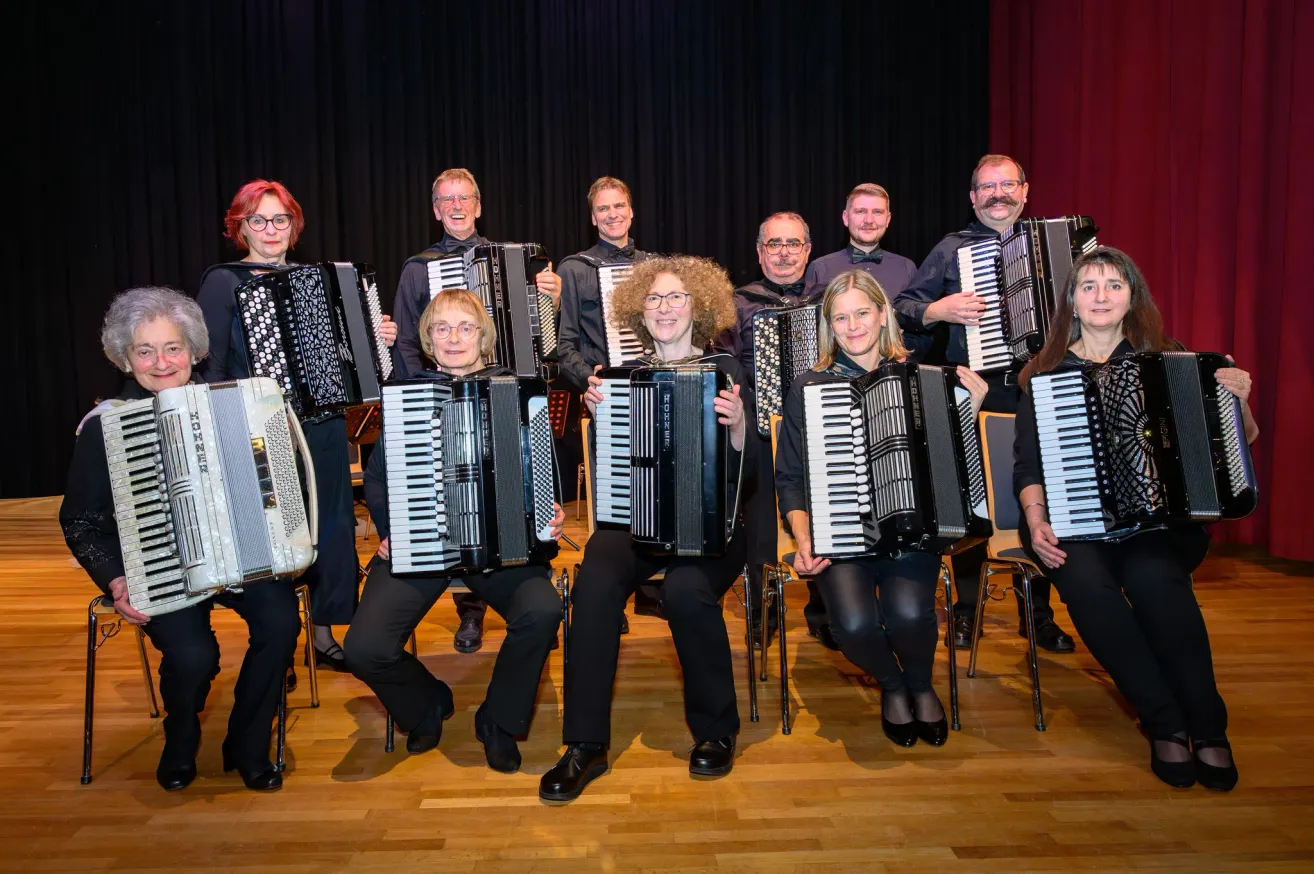 A group of musicians are posing for a photo, all holding accordions. They are seated and standing, smiling, against a black curtain backdrop.