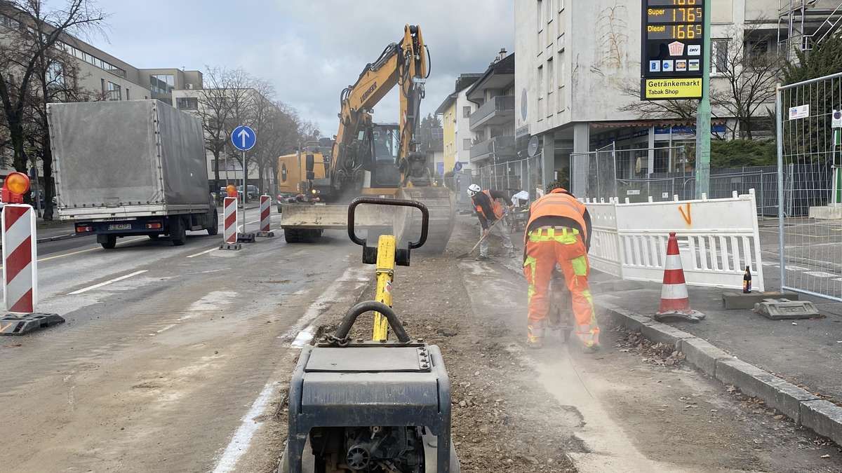 Construction workers are working on the sidewalk, with a yellow excavator nearby, and a truck parked on the road. The road has white markings, and a traffic sign is on the left. Buildings are on the right, and a signboard with the text 'Getrunken Tabak Shop' is mounted on the wall.