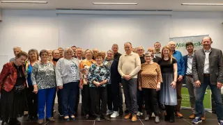 A group of people standing together for a photo. They are all wearing casual clothes. The man in the middle is wearing a polo shirt and leather shoes. The woman on the right is wearing a dress and boots. Behind them is a white wall with a green sign on it. The floor is black and shiny.