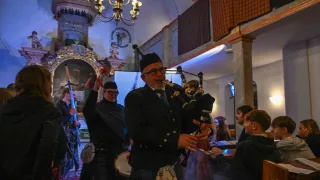 A bagpiper in a kilt plays music in a church with a chandelier. A man in a suit watches and a crowd watches.
