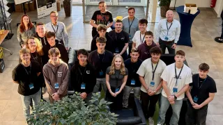 A group of people, likely students, pose for a photo with a woman in the middle. They wear IDs and some wear glasses. They stand in front of a potted plant, with a couch and glass door behind them.