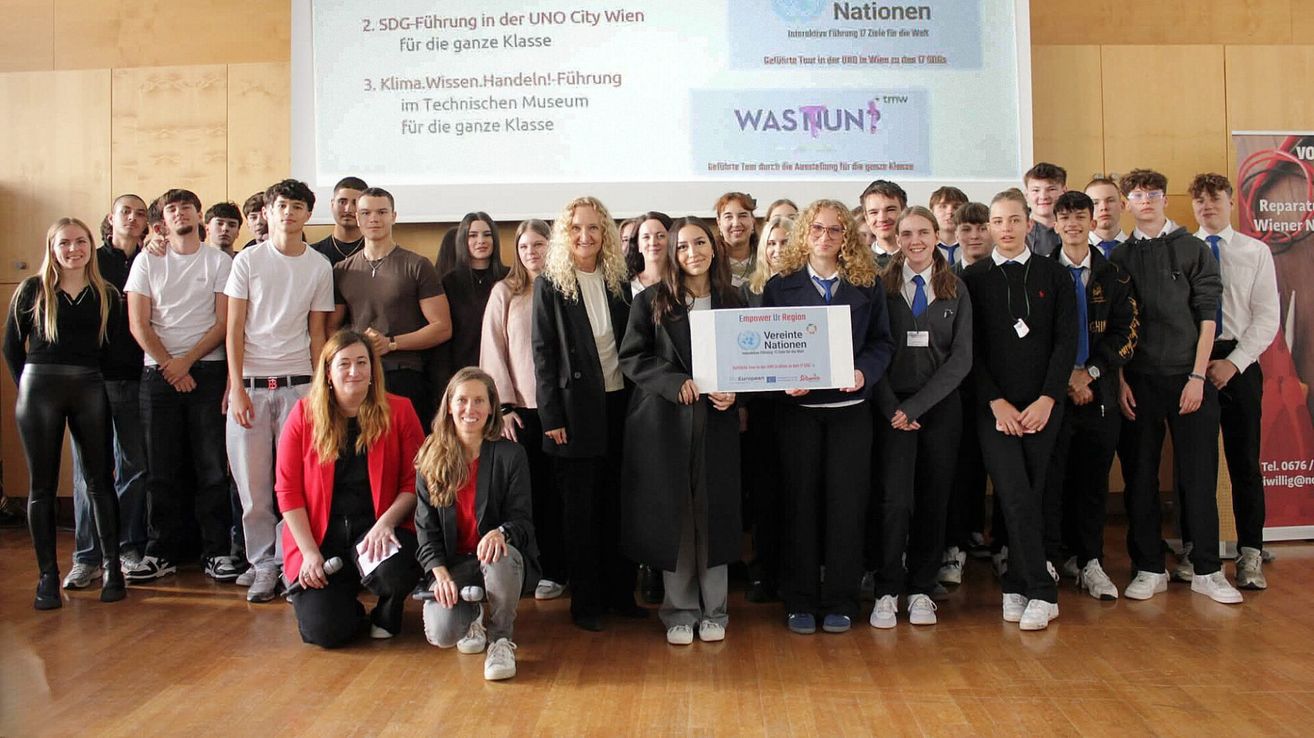 A group of students stands with two women, one holding a certificate, in front of a presentation backdrop with SDGs and climate action topics.