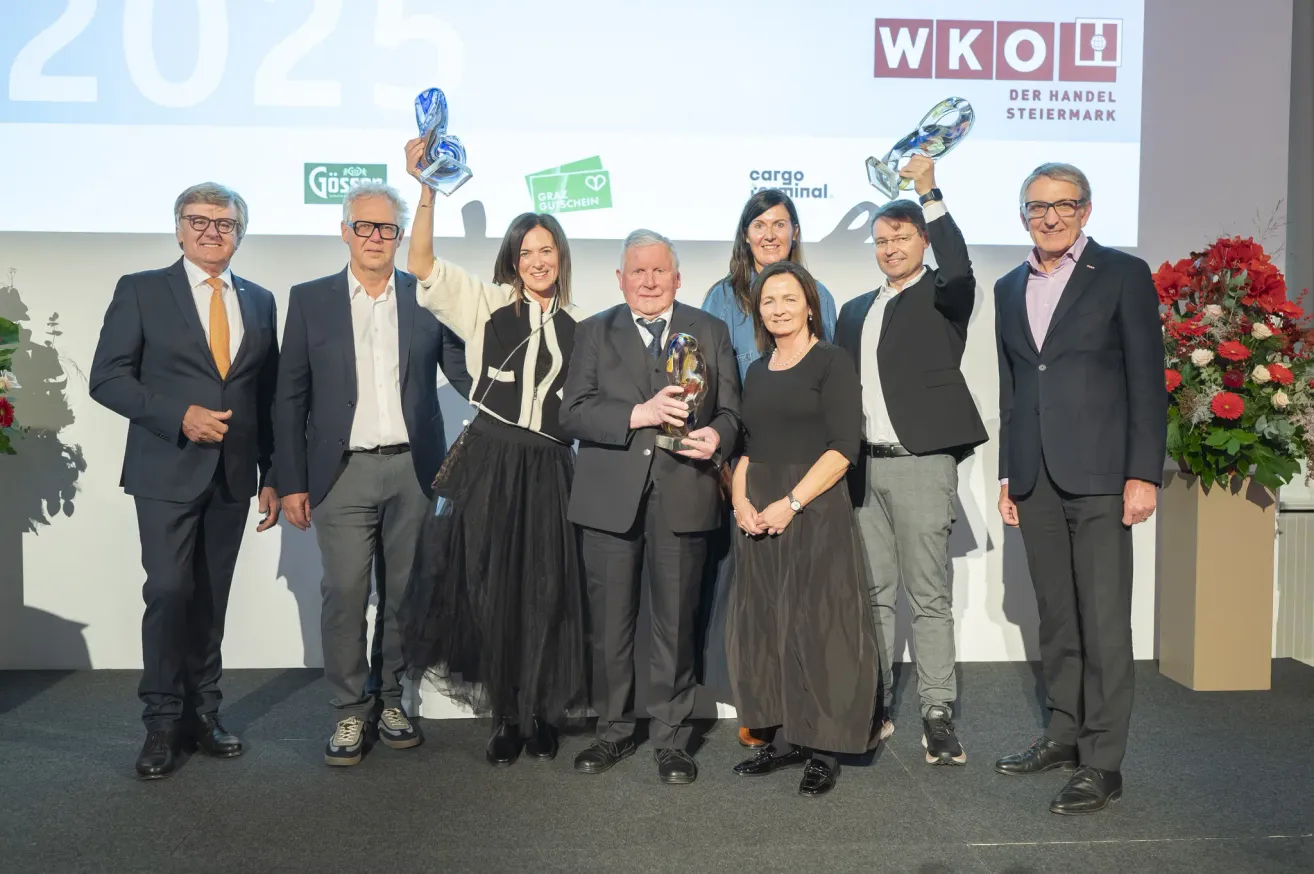 A group of people dressed in formal attire stand on a stage, holding awards. Behind them, a banner with the text 'WKO' and 'DER HAND STEIRMARK' is visible. The individuals seem to be posing for a photograph.