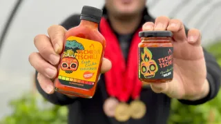 A man holding two bottles of TSCHIDA hot sauce, one large and one small, both with vibrant labels. He stands in a greenhouse, wearing a medal around his neck.