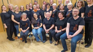 A group of people wearing black t-shirts with orange text are sitting on chairs in a room. They are smiling and posing for a photo.