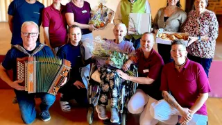 A group of people are gathered, smiling and posing for a photo. A woman in a wheelchair is at the center, holding flowers. Others hold gifts and musical instruments. The setting is indoors with wooden floors.