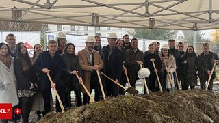 A group of people wearing winter attire and helmets gather under a tent to break ground for a construction project. They hold wooden shovels and pose for a photo.