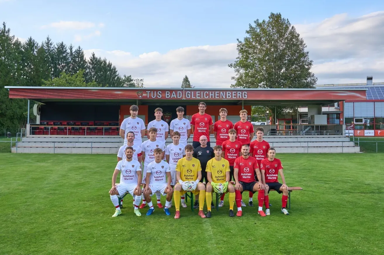 A soccer team in red and white uniforms poses for a photo on a green field with a red building labeled 'Tus Bad Gleichenberg' in the background.