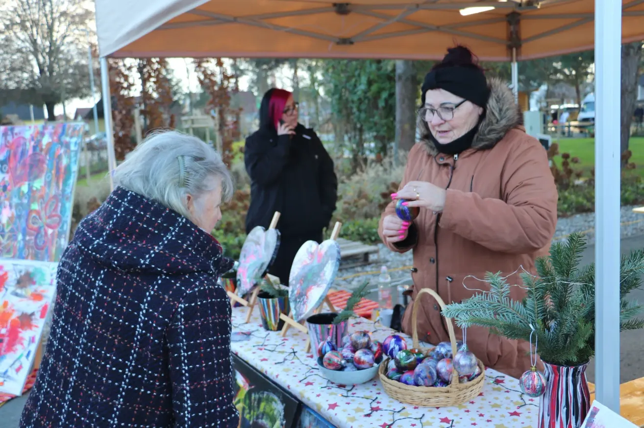 Three women stand under a tent at an outdoor market. One woman holds an ornament, another talks on her phone, and the third looks at ornaments on a table.