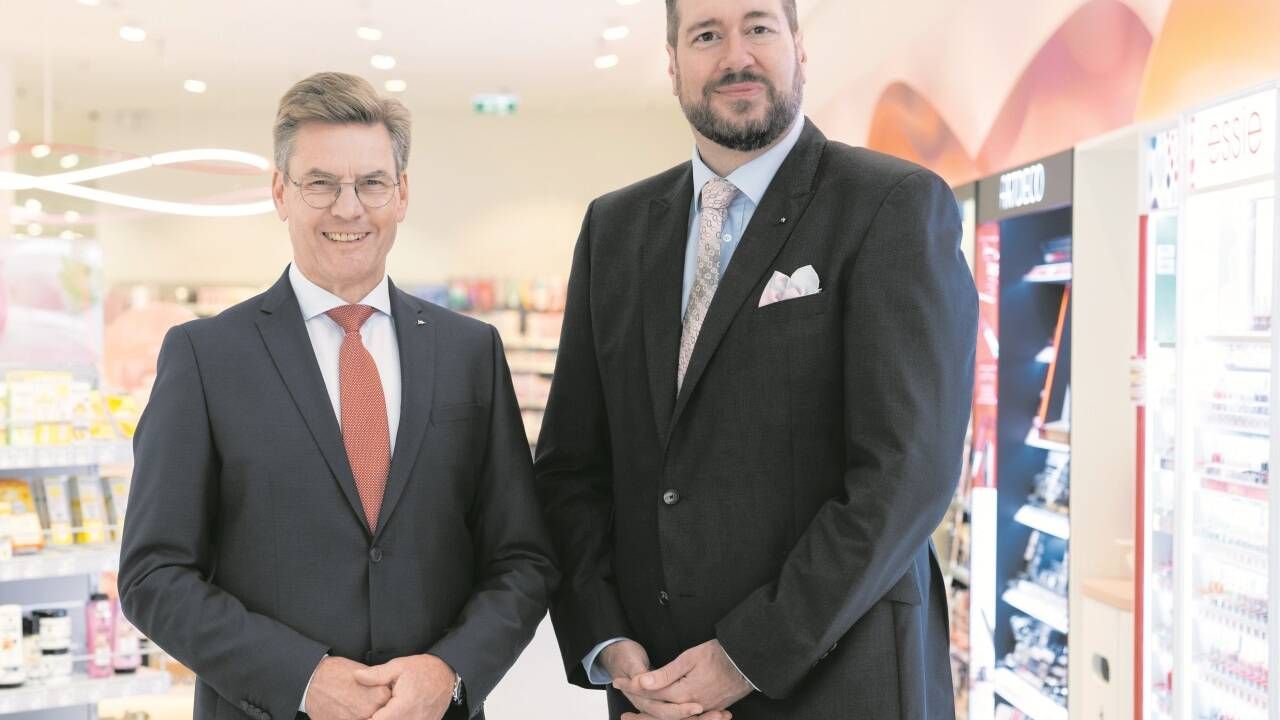 Two men in suits stand in a store, one smiling, the other with a beard. They are standing next to each other.