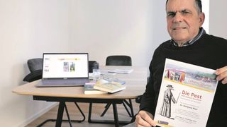 An older man smiles while holding a book about the plague. A laptop is open on the table in front of him.